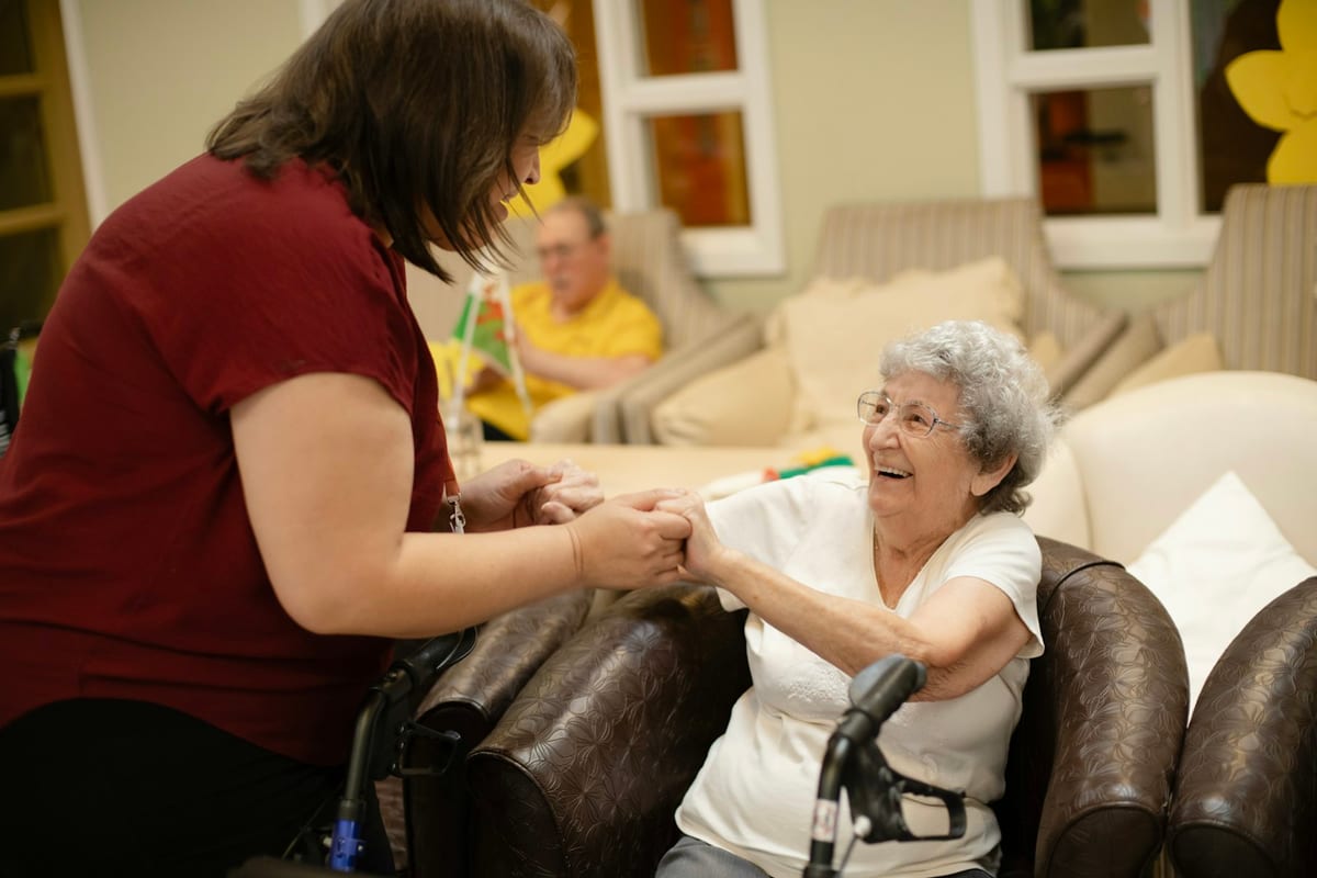 A caregiver holding hands with an older woman seated beside her walker